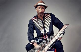 Image shows a man wearing a dark blue top with black and white stripes sat on a chair and holding an instrument.