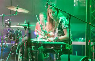 Image shows a girl on stage playing the drums with a microphone next to her.