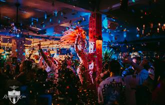 Photo of a crowded bar with fantastic dancing performers in feather headpieces, glitter faling from the ceiling and disco lights.