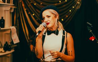 Image of young lady with blonde hair wearing a beret, bow tie and braces performing a spoken word piece on stage.