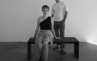 Image of a lady sat on a bench with a young man stood behind.