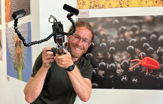 Alex Hyde holding a camera and smiling, with the sherwood forest up close exhibition wall behind