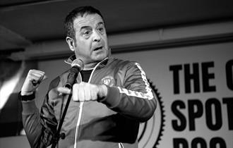 Image shows a black and white image of Mark Thomas on stage during his show - Gaffa Tapes.