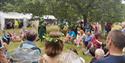 A crowd sits in a circle on a field for the Sherwood Festival. They sit engaged in a story by a cosumted figure.