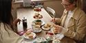 Two females, one brunette and one blonde with glasses are enjoying an Afternoon Tea serving for two. The blonde female is spreading jam on her scone.