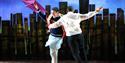 The Kite Runner – Two men dance with opposite arms around each other’s waists. One holds a purple kite and smiles. In the background is the shape of a