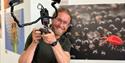 Alex Hyde holding a camera and smiling, with the sherwood forest up close exhibition wall behind