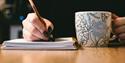 A hand holding a pencil writing on a notepad with a mug next to it