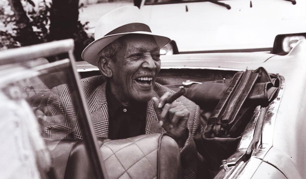 Edgar Macías Quintet, smiling, smoking cigar in a covertible car.