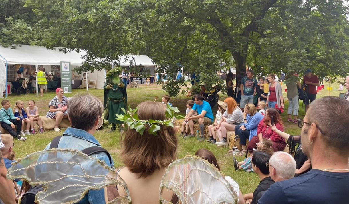 A crowd sits in a circle on a field for the Sherwood Festival. They sit engaged in a story by a cosumted figure.