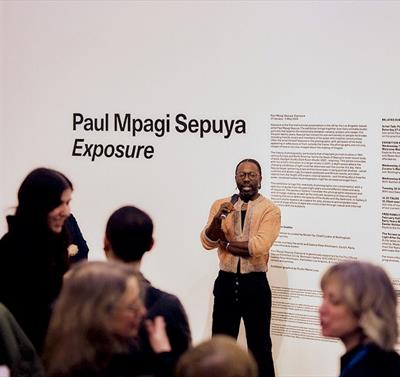 Photo of Paul Mpagi Sepuya on a walkthrough event. He is holding a microphone and you can see his gallery at Nottingham Contemporary in the background