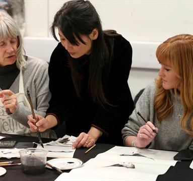 Photo of Feng-Ru Lee during a workshop. She is leaning over and assisting two women, who are holding paint brushes.