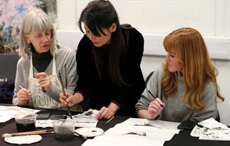 Photo of Feng-Ru Lee during a workshop. She is leaning over and assisting two women, who are holding paint brushes.