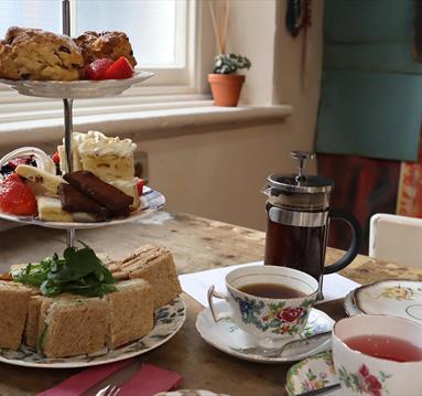A serving of Afternoon Tea for two. A three tier cake stand showcasing sandwich squares, a mixture of cakes and fruit scones. Two teacups and saucers 