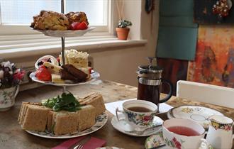A serving of Afternoon Tea for two. A three tier cake stand showcasing sandwich squares, a mixture of cakes and fruit scones. Two teacups and saucers