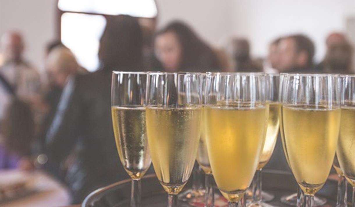 The image shows a circular tray of glasses filled with prosecco with a blurred background of people sat theatre style