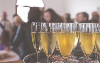 The image shows a circular tray of glasses filled with prosecco with a blurred background of people sat theatre style
