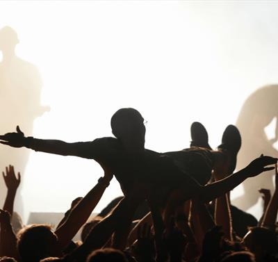 Photo of crowd surfer. They are in silhouette against bright white stage lights in the background. You can see some performers on stage.