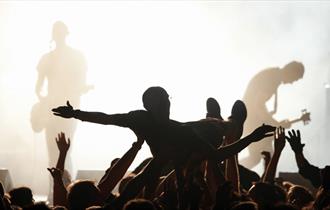 Photo of crowd surfer. They are in silhouette against bright white stage lights in the background. You can see some performers on stage.
