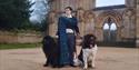 An actor dressed as Lord Byron poses in front of Newstead Abbey with two large Newfoundland dogs.