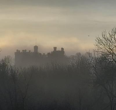 A photograph of Belvoir Castle with an atmospheric ambience. Fog shrouds the castle, which you can only just make out in the background, silhouetted a