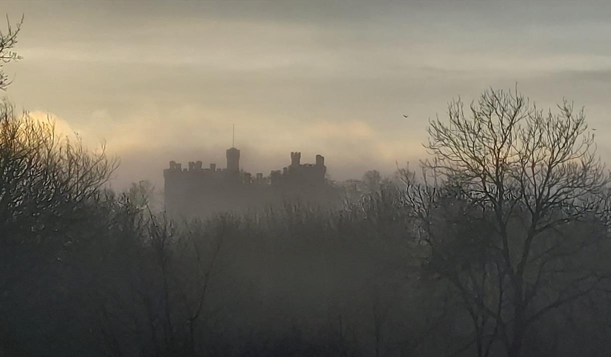 A photograph of Belvoir Castle with an atmospheric ambience. Fog shrouds the castle, which you can only just make out in the background, silhouetted a