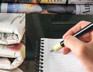 A hand holding a pen writing in a notebook next to a stack of newspapers