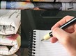A hand holding a pen writing in a notebook next to a stack of newspapers