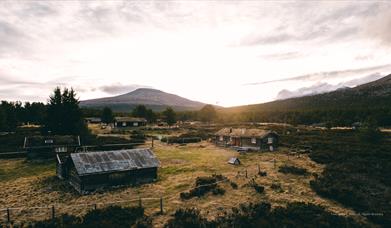 Utsikt over Mistdalen i Rendalen en stille kveld, med lav sol som skinner gjennom skyene og belyser det åpne landskapet. Tradisjonelle trehytter med torvtak ligger spredt i terrenget, omgitt av frodig skog og majestetiske fjell i bakgrunnen. Elven slynger seg gjennom dalen, og området utstråler en rolig, uberørt villmarksstemning.