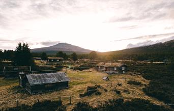 Utsikt over Mistdalen i Rendalen en stille kveld, med lav sol som skinner gjennom skyene og belyser det åpne landskapet. Tradisjonelle trehytter med torvtak ligger spredt i terrenget, omgitt av frodig skog og majestetiske fjell i bakgrunnen. Elven slynger seg gjennom dalen, og området utstråler en rolig, uberørt villmarksstemning.