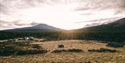 Utsikt over Mistdalen i Rendalen en stille kveld, med lav sol som skinner gjennom skyene og belyser det åpne landskapet. Tradisjonelle trehytter med torvtak ligger spredt i terrenget, omgitt av frodig skog og majestetiske fjell i bakgrunnen. Elven slynger seg gjennom dalen, og området utstråler en rolig, uberørt villmarksstemning.