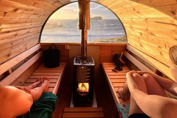 2 people sitting inside a barrel sauna looking out to the sea
