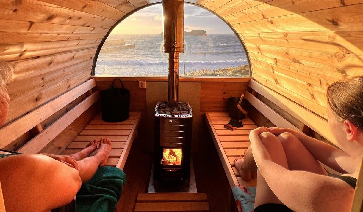 2 people sitting inside a barrel sauna looking out to the sea