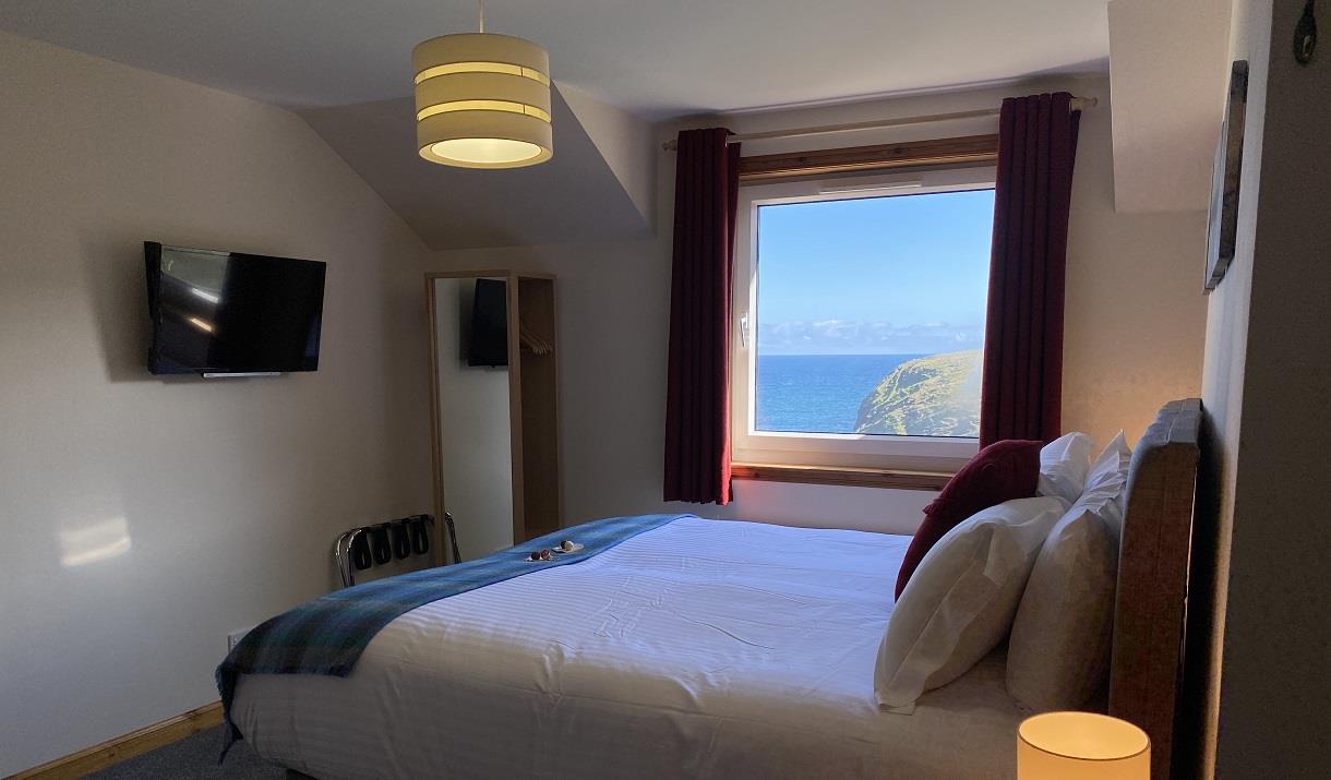 A super king size bed with fresh white linen and colourful tweeds in front of a window overlooking the ocean.