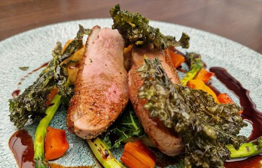 Pan fried duck breast with roast vegetables and golden jus sitting on a speckled blue plate.