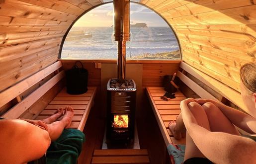 2 people sitting inside a barrel sauna looking out to the sea