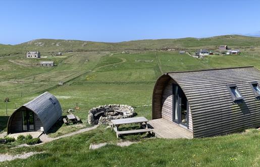 Two curved wooden cabins with picnic tables sit in a grassy meadow with a circular stone fire pit between them.