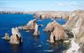 A view of the Mangersta Sea Stacks, sharp jagged rocks standing in a calm blue ocean. The Mangersta sea cliffs stretch out behind them.