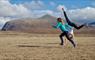 Two girls are running and turning cartwheels on a flat grassy plain with Mealaisabhal, a rocky hill with a conical peak behind them.