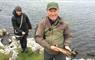 A smiling man and woman, dressed in outdoor gear are standing next to a loch. The man is holding a brown trout and the woman a fishing rod.