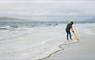 Photo of an artist on a sandy beach, drawing a line round the tide line with an oversized pencil.