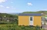 A yellow Shepherd's Hut in a wild flower meadow. Behind it is a stone wall and grassy hillside leading to the ocean.