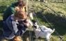 A young blonde couple are crouching down next to a fence, bottle feeding two blackface lambs.