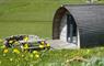 Curved wooden clad cabin at Mangersta Croft Holidays, in green croft fields covered in yellow flowers. Decking, a picnic bench and stone fire pit are
