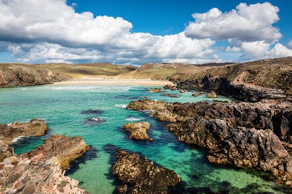 Rocky coastline with clear turquoise waters and sandy beach under a bright blue sky with scattered clouds. Rugged terrain surrounds the water.