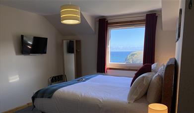 A super king size bed with fresh white linen and colourful tweeds in front of a window overlooking the ocean.