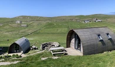 Two curved wooden cabins with picnic tables sit in a grassy meadow with a circular stone fire pit between them.