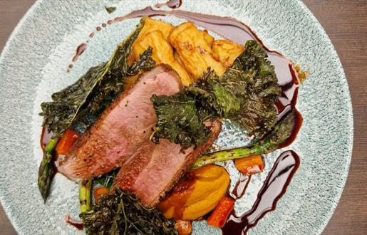 Duck breast with roasted vegetables and sauce on a blue plate, sitting on a brown table.