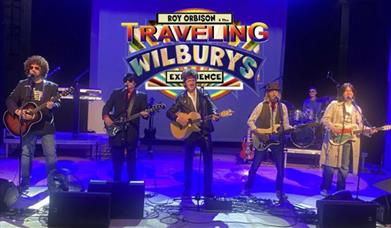 Roy Orbison & the Travelling Wilbury's performing on stage under blue lighting.