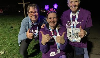Three people smile and give thumbs up wearing Starlight Hike t-shirts, medals and flashing headwear, outdoors at Starlight Hike in Ferry Meadows.